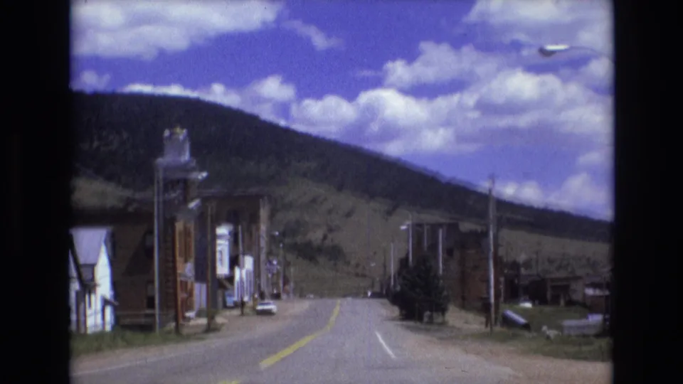 1973 Colorado-area 8mm frame showing a curving town road with small buildings, a streetlamp, utility poles, surrounding hills, and a bright clouded sky.