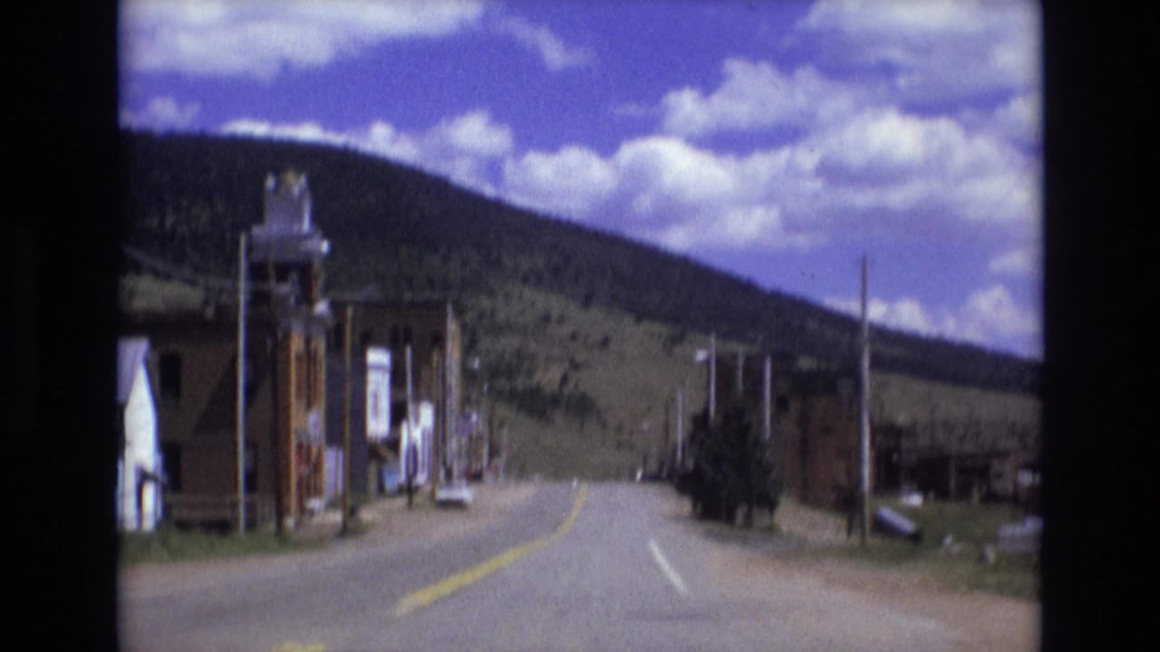1973 8mm frame from Colorado Springs showing a quiet paved street, brick buildings, utility poles, and a forested hill under scattered clouds.