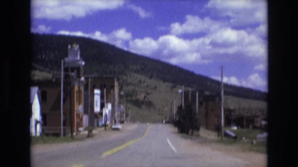 1973 8mm frame from Colorado Springs showing a quiet paved street, brick buildings, utility poles, and a forested hill under scattered clouds.