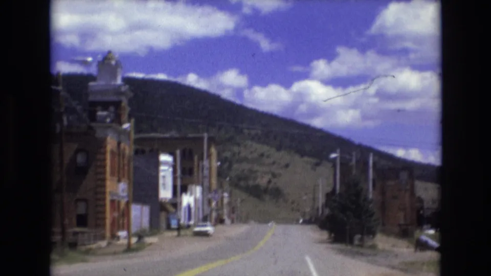 Moving 8mm view along a Colorado Springs street with brick facades close to the camera, a parked car, utility poles, and the hill rising behind town.