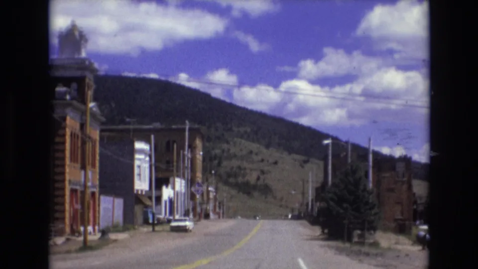 Late-frame view from the 1973 Colorado Springs drive showing brick commercial buildings, a quiet paved street, evergreens, utility poles, and a broad hillside beneath clouds.
