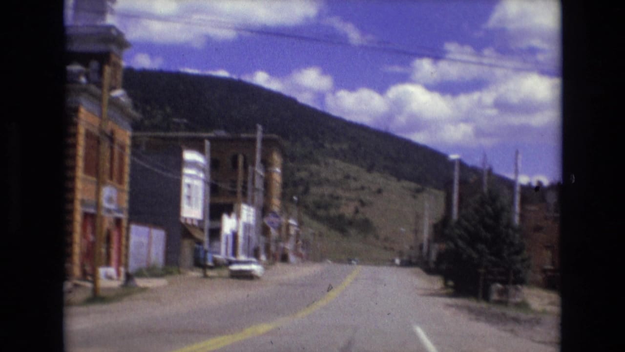1973 Colorado Springs area home-movie frame showing a small-town street bordered by brick buildings with a hillside rising behind it.