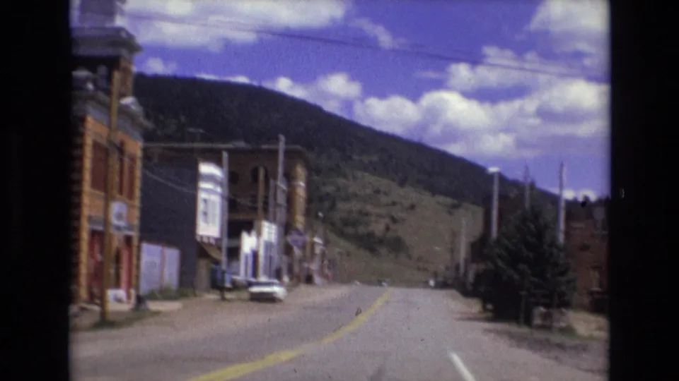 1973 Colorado Springs area home-movie frame showing a small-town street bordered by brick buildings with a hillside rising behind it.