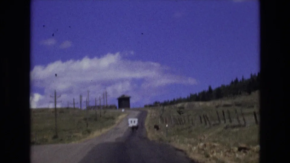 1973 Colorado countryside view from the road, with a paved lane rising through open fields toward a small dark roadside or hilltop structure.