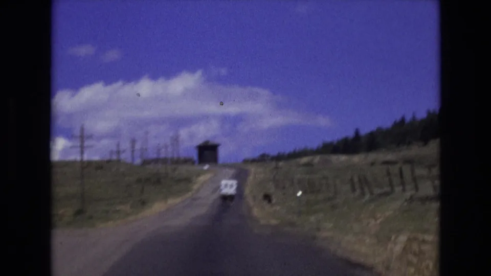 1973 Colorado rural driving frame with a white vehicle ahead on a paved road bordered by fields, fence posts, and utility poles.