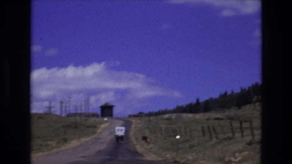 1973 Colorado countryside frame showing a white vehicle traveling uphill on a narrow paved road with fenced fields and a small structure in the distance.