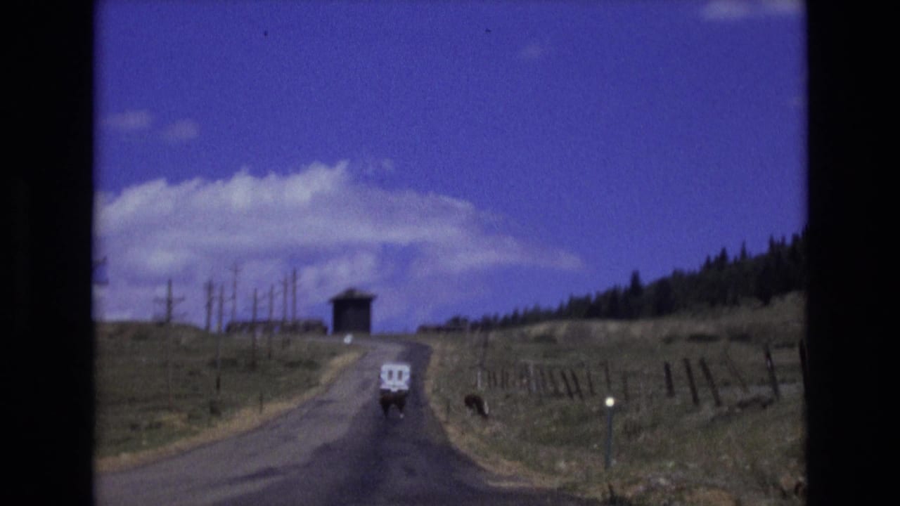 1973 Colorado Springs 8mm frame showing a white van traveling away on a quiet rural road between grassy hills, fence posts, telephone poles, and a small dark structure in the distance.
