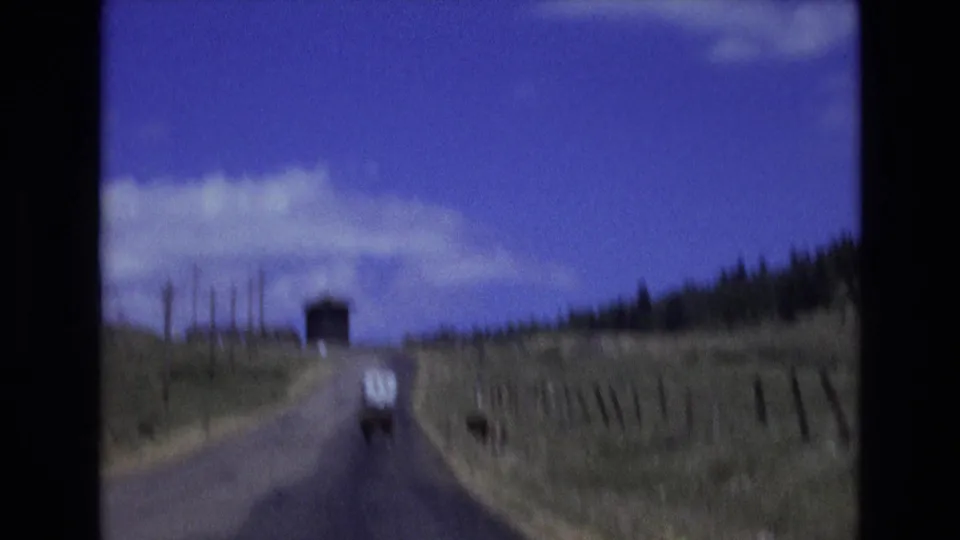 1973 Colorado Springs home-movie frame showing a white van moving along a curving rural road past grassland, fence posts, telephone poles, a tree line, and a small dark structure ahead.