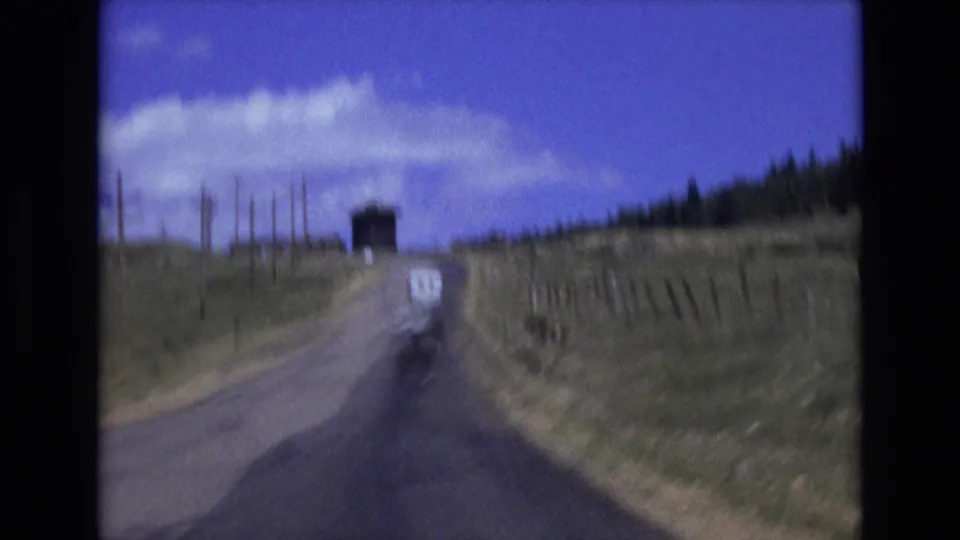 Final 8mm frame from the Colorado Springs road sequence, with the white van centered ahead on the pavement and open hills, fence posts, clouds, and a dark tree line framing the route.