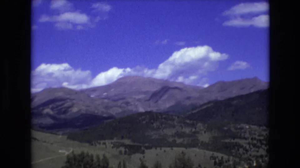 1973 8mm landscape frame near Colorado Springs showing a broad mountain valley, dark forested slopes, open green land, and scattered white clouds in a blue sky.