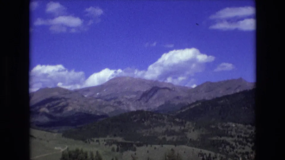 1973 Colorado Springs-area home-movie frame of distant mountain ridges, a green valley floor, dark tree-covered slopes, and bright sky with small clouds.