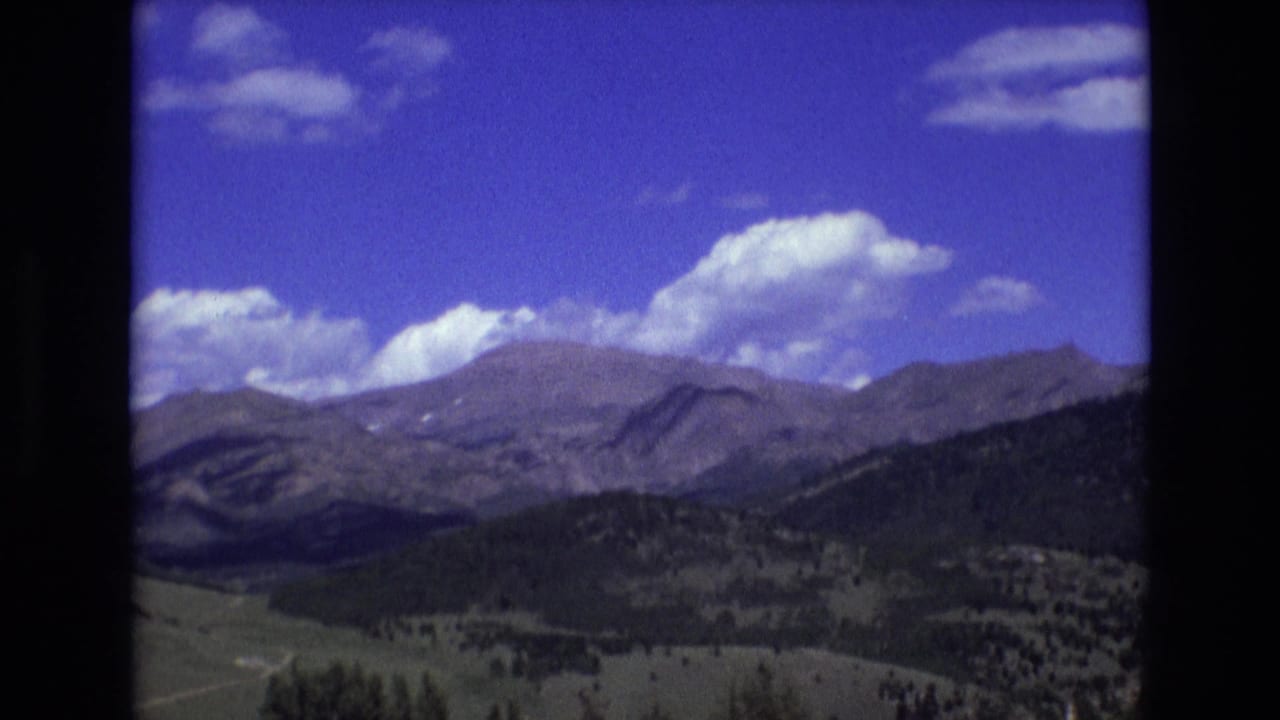 1973 8mm home-movie frame of a Rocky Mountain landscape near Colorado Springs, with distant ridges, dark forested slopes, open valley land, and scattered clouds in a blue sky.