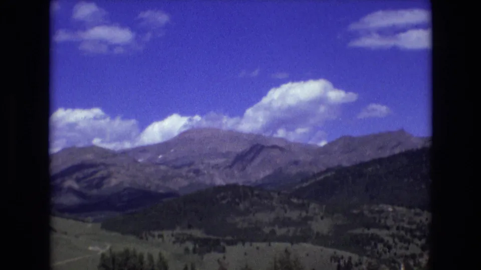 1973 8mm home-movie frame of a Rocky Mountain landscape near Colorado Springs, with distant ridges, dark forested slopes, open valley land, and scattered clouds in a blue sky.
