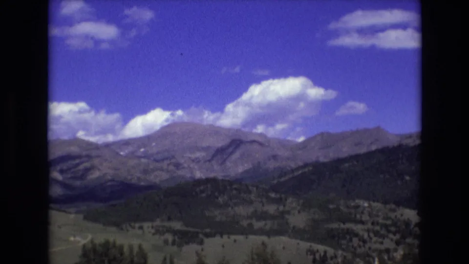 1973 8mm travel footage frame from the Colorado Springs area, with a soft mountain panorama, darker tree-covered slopes, open green land, and large white clouds across the sky.