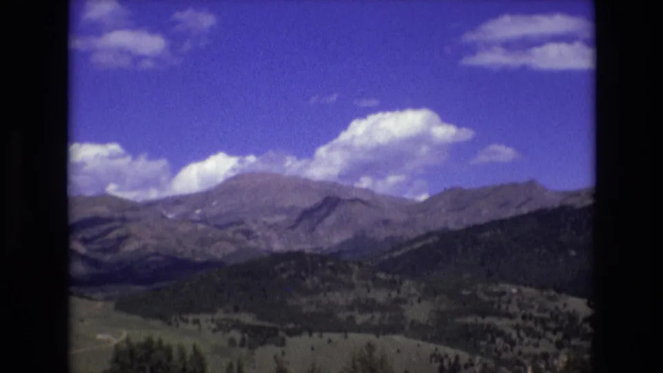 1973 8mm home-movie frame of the Rocky Mountain landscape near Colorado Springs, showing forested hills in the foreground, distant ridges, valley terrain, and scattered white clouds.