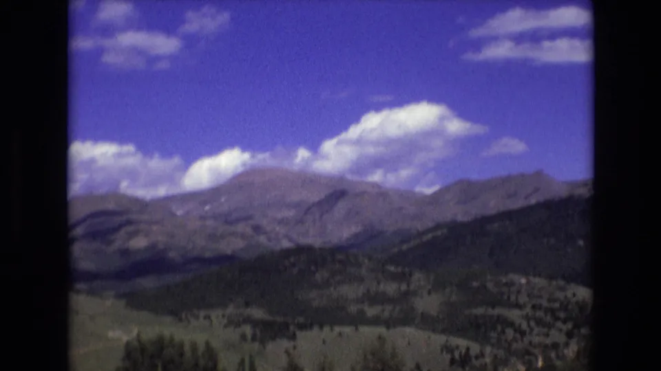 1973 Colorado Springs 8mm frame showing distant rocky mountains, dark evergreen foothills, open grassland, and scattered white clouds in a saturated blue sky.