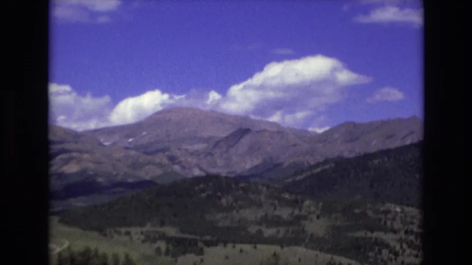 1973 Colorado Springs 8mm frame with a broad rocky ridge, shadowed evergreen hills, green foothill patches, and bright cumulus clouds overhead.