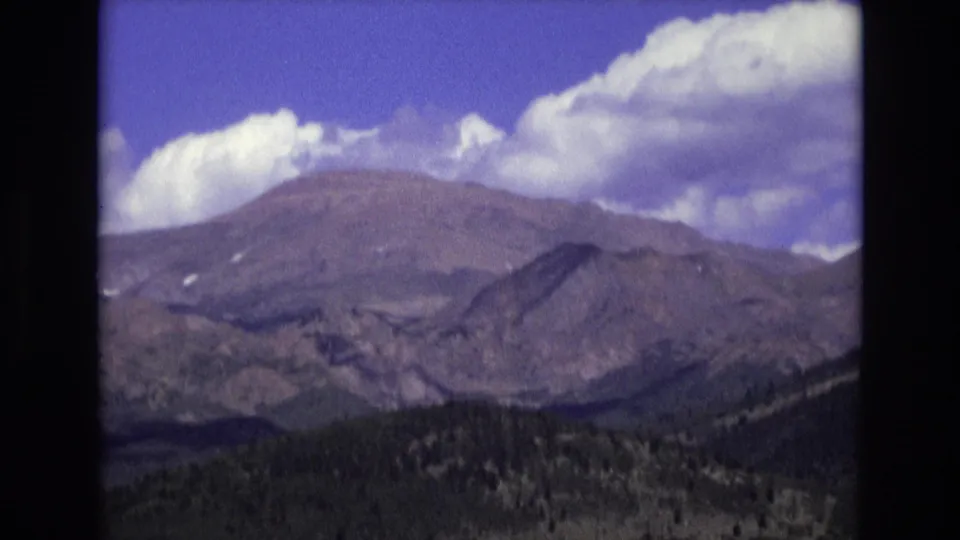 1973 Colorado Springs 8mm frame showing a closer view of brown mountain slopes, heavy white clouds, dark tree-covered foothills, and soft home-movie focus.