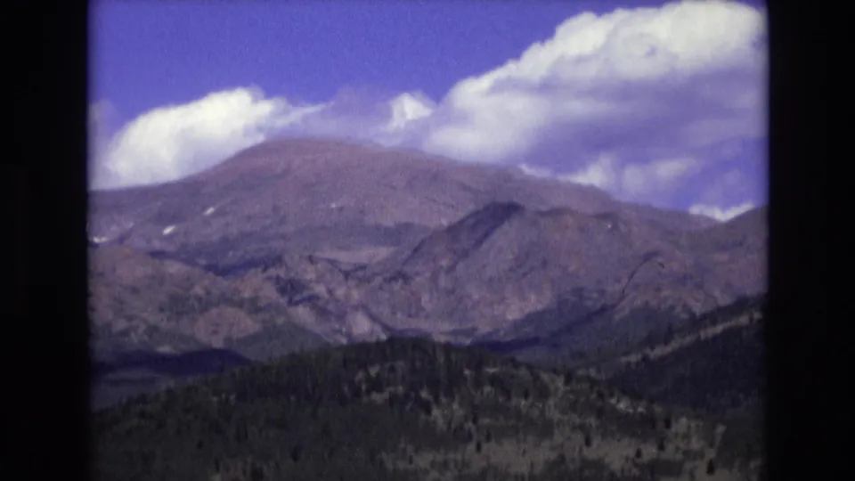 1973 Colorado Springs 8mm frame showing rugged mountain slopes, a dark forested valley, scattered green clearings, and a large cloud bank across the upper sky.