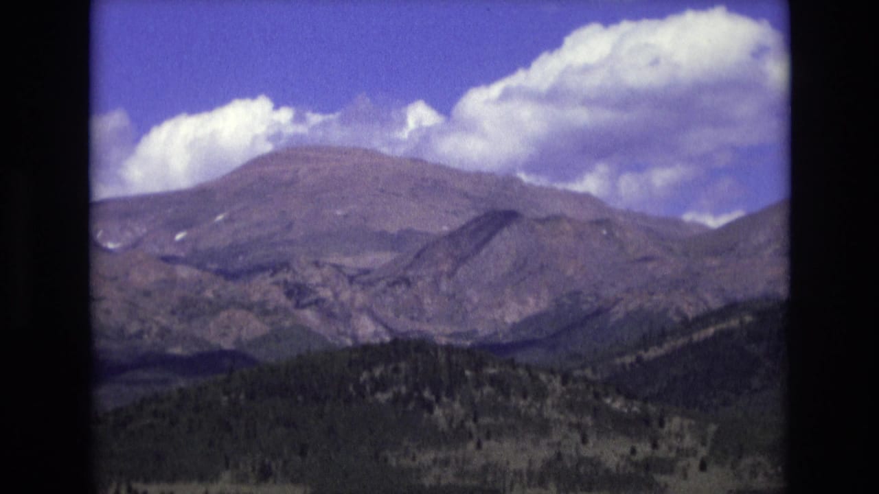 1973 8mm home-movie frame from Colorado Springs showing a broad mountain ridge, dark foothills, and white clouds against a saturated blue sky.