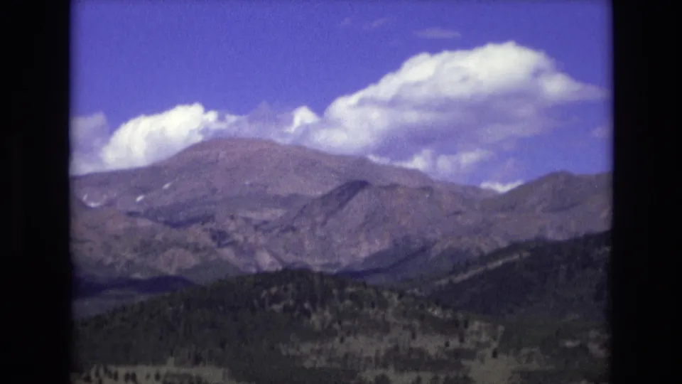 1973 Colorado Springs landscape frame with rocky mountain slopes, shadowed foothills, and large cumulus clouds in a bright blue sky.