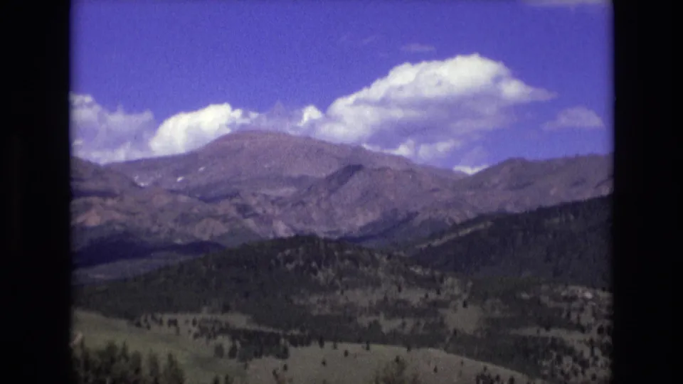 1973 8mm Colorado Springs frame showing a wider mountain and valley view with green meadows, scattered trees, and clouds over the ridge line.