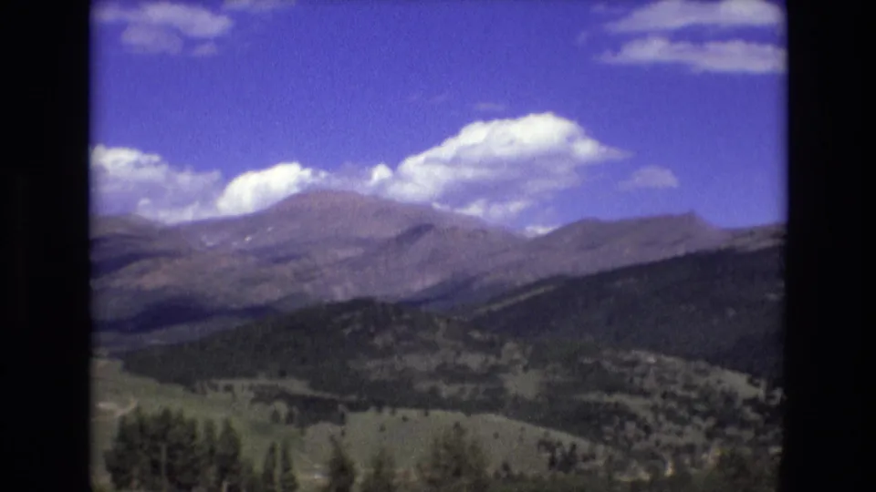 Wide 1973 Colorado Springs home-movie frame showing green hills and dark tree cover leading toward a mountain range beneath bright white clouds.