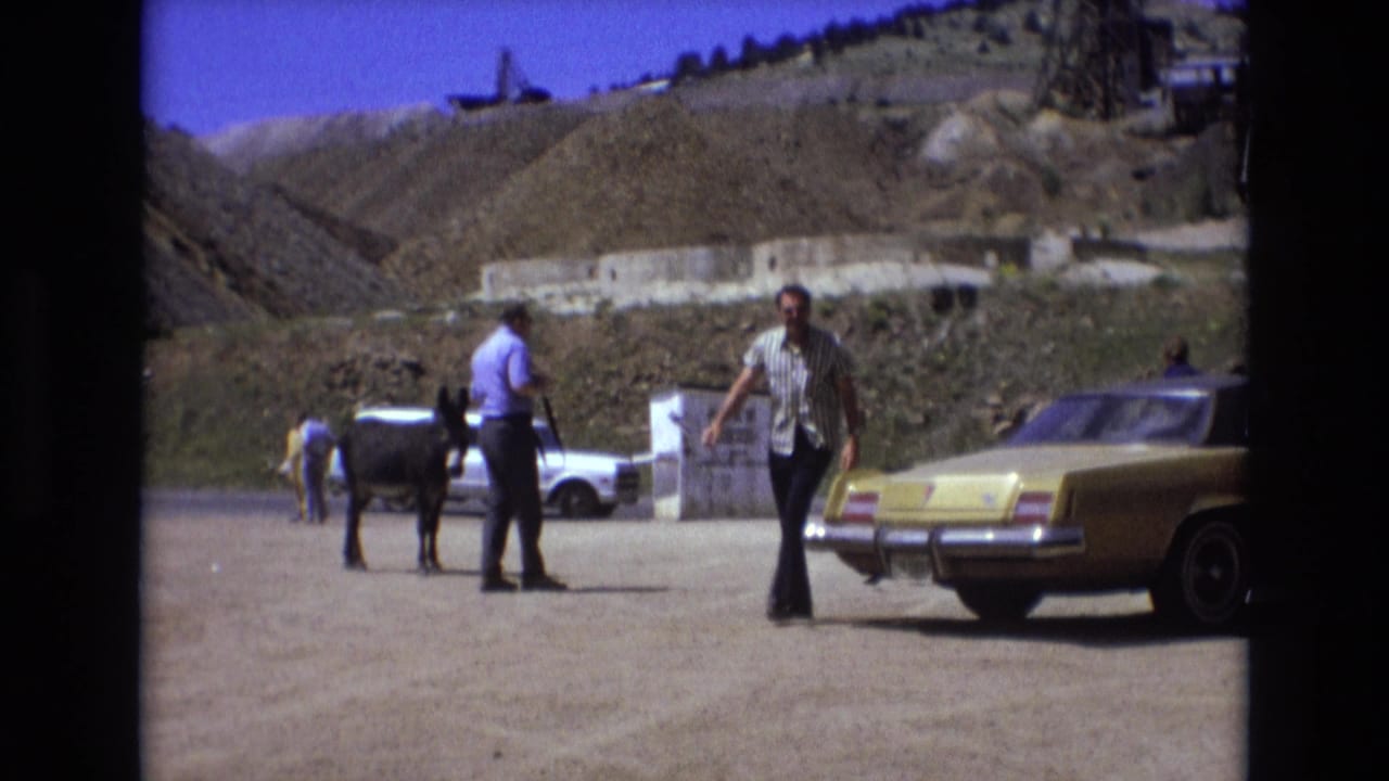 A 1970s roadside scene in the supplied Colorado Springs location shows a man walking past a yellow sedan as another adult stands near a donkey, with hills and structures behind them.