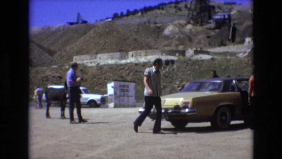 A man in striped clothing crosses the foreground between a donkey and a yellow 1970s sedan at a dusty roadside attraction in the Colorado Springs footage.