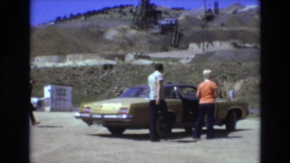 Two adults stand near an open door of a yellow sedan in the supplied 1973 Colorado Springs home-movie footage, with excavated hills and roadside structures in the background.