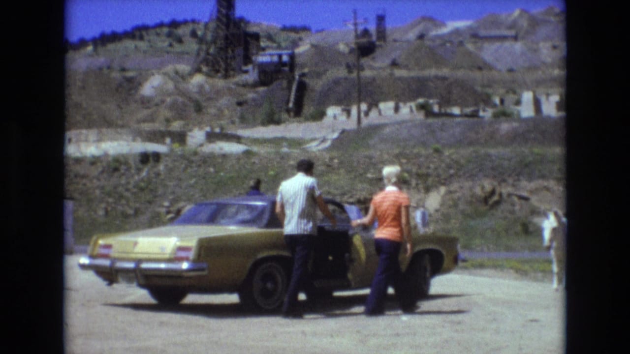 In 1973 Colorado Springs home-movie footage, a man and woman stand beside a gold-colored sedan on a dusty roadside while a white donkey stands near arid hills and industrial structures.