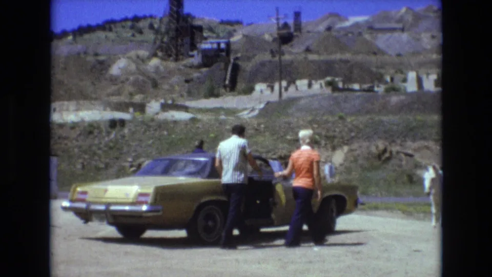 In 1973 Colorado Springs home-movie footage, a man and woman stand beside a gold-colored sedan on a dusty roadside while a white donkey stands near arid hills and industrial structures.