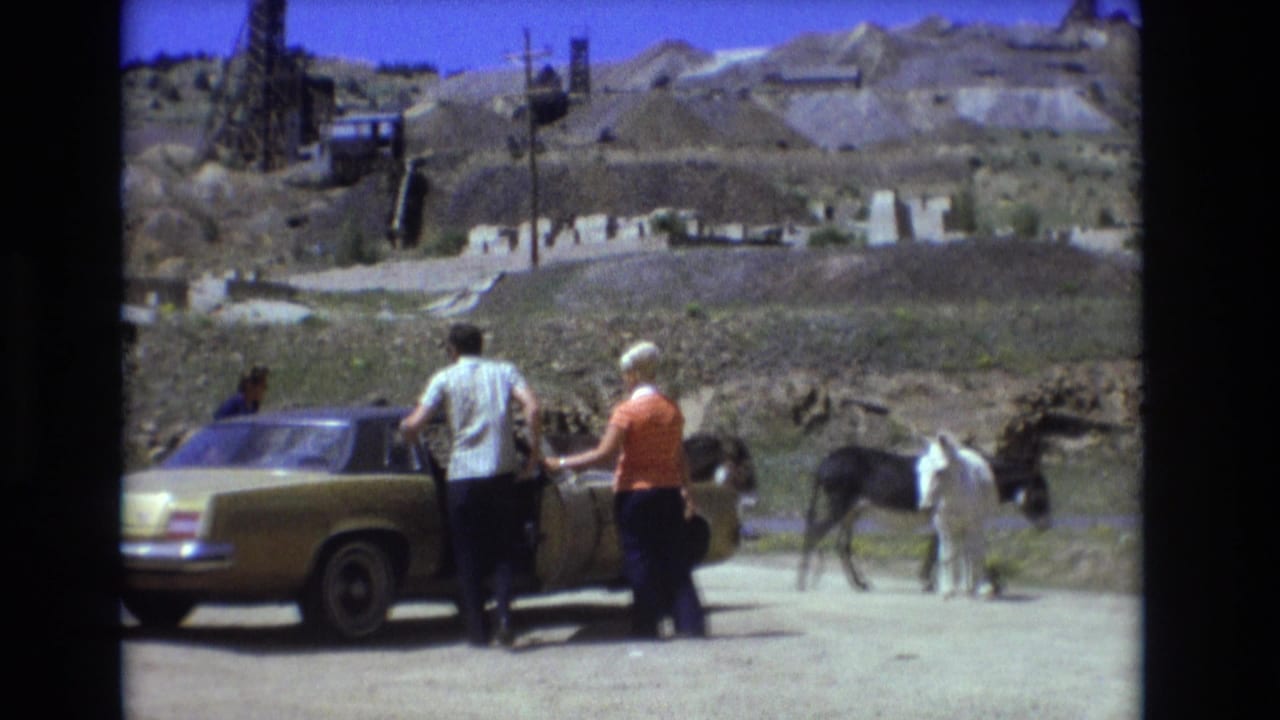 In metadata-supported 1973 Colorado Springs footage, adults stand by a gold sedan as donkeys graze near a dusty roadside with an excavated hillside and dark structures behind them.