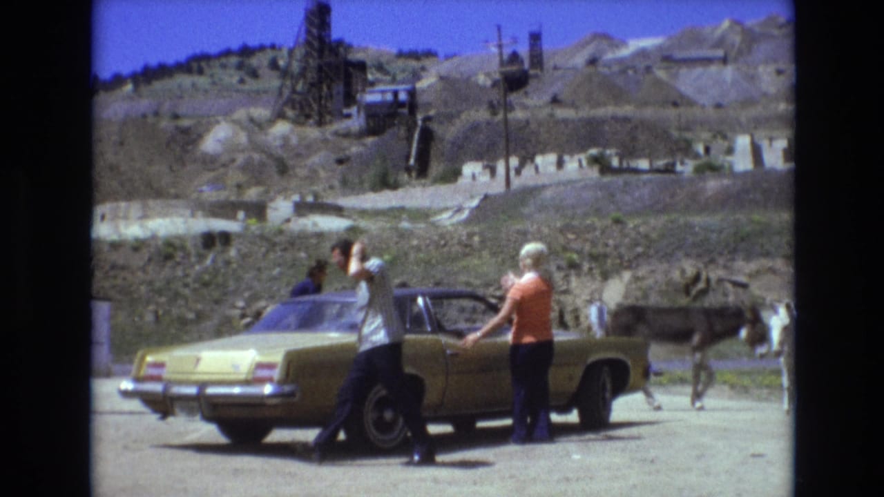 In the 1973 Colorado Springs home-movie scene, a man walks from a gold sedan while a woman remains by the car and donkeys pass along the dusty roadside below the hillside.