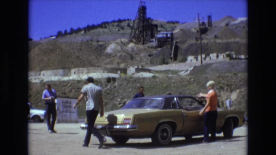 A man crosses the dusty road near a parked gold sedan while a woman stands at the passenger side, with Colorado Springs hillside excavation and roadside structures visible in the background.