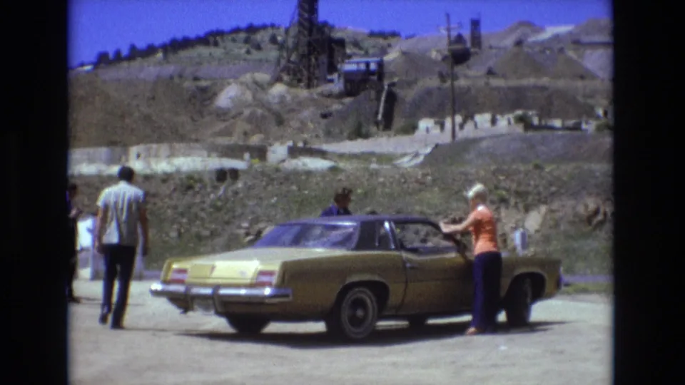 The 1973 Colorado Springs home-movie frame shows a woman beside a gold sedan, a man walking away, and donkeys near the roadside beneath a bright sky and excavated hillside.