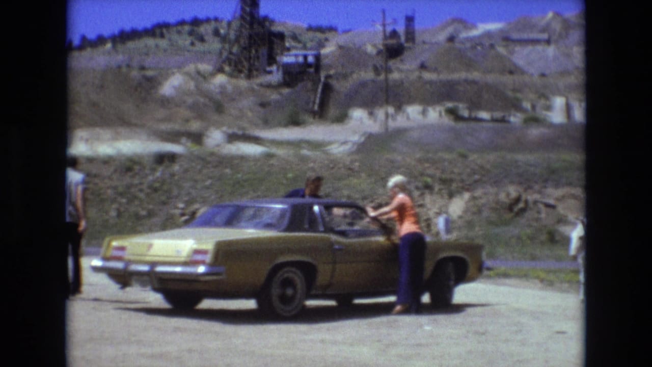 1973 Colorado home-movie frame showing a gold sedan stopped near Battle Mountain, with an adult woman in an orange top by the car and mining structures in the arid background.