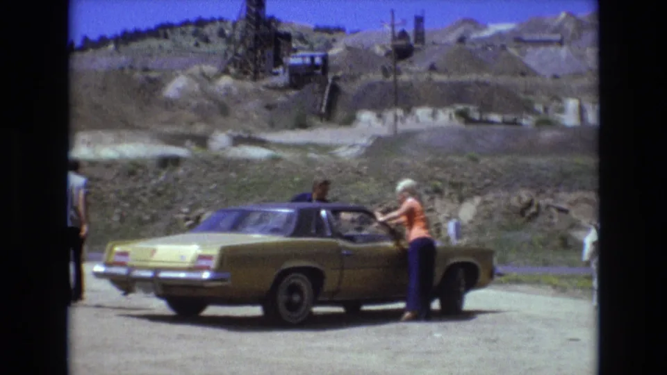 1973 Colorado home-movie frame showing a gold sedan stopped near Battle Mountain, with an adult woman in an orange top by the car and mining structures in the arid background.