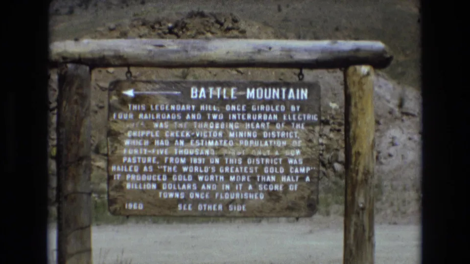 Battle Mountain historical marker in Colorado, filmed close enough to show the title and mining-history text, with rough timber supports and dry hillside behind it.