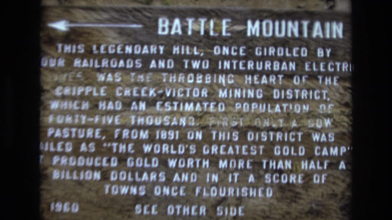 1973 Colorado 8mm frame showing a close view of the Battle Mountain historical marker, with white text describing the Cripple Creek-Victor Mining District on a dark wooden sign.