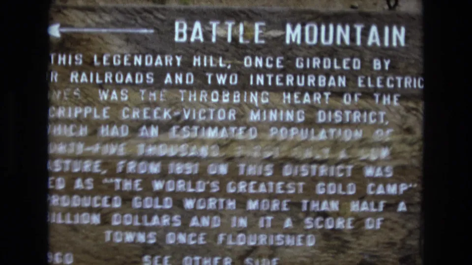 Close 8mm view of the Battle Mountain sign in Colorado, with weathered wood grain and text mentioning the Cripple Creek-Victor Mining District.