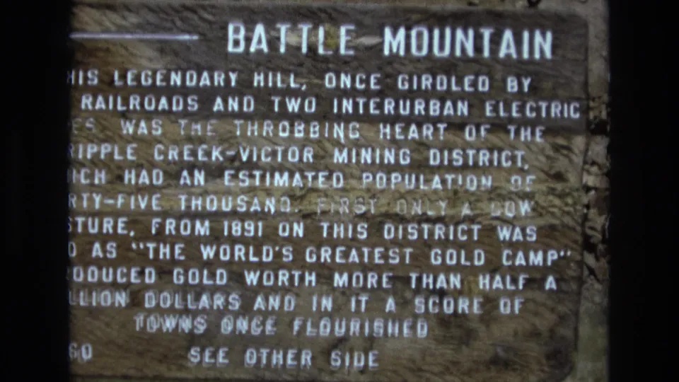 Close frame of the Battle Mountain roadside marker, showing its mining-history text and weathered wooden face in 1970s 8mm footage.