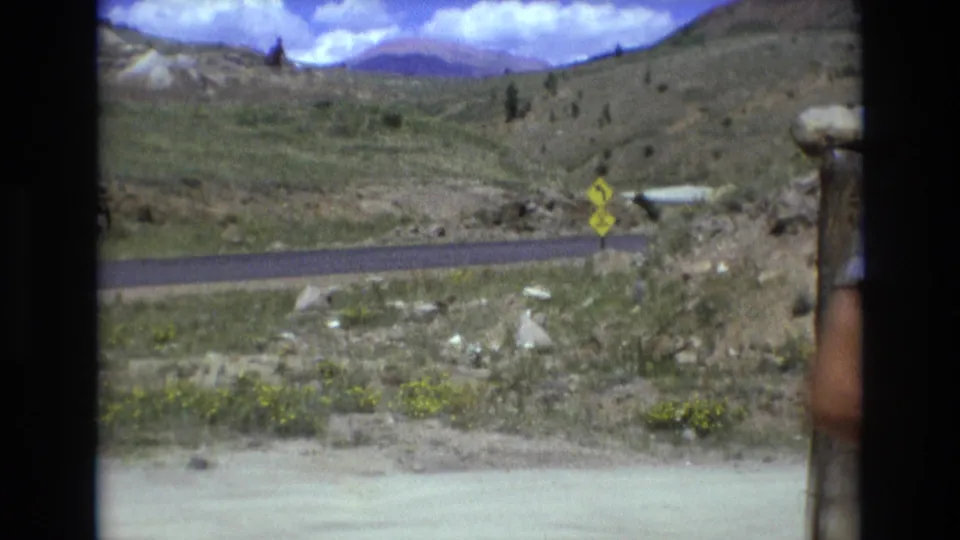 1973 Colorado foothills landscape with a paved road, roadside sign, grassy slopes, rocky ground, and blue sky after the Battle Mountain marker shot.