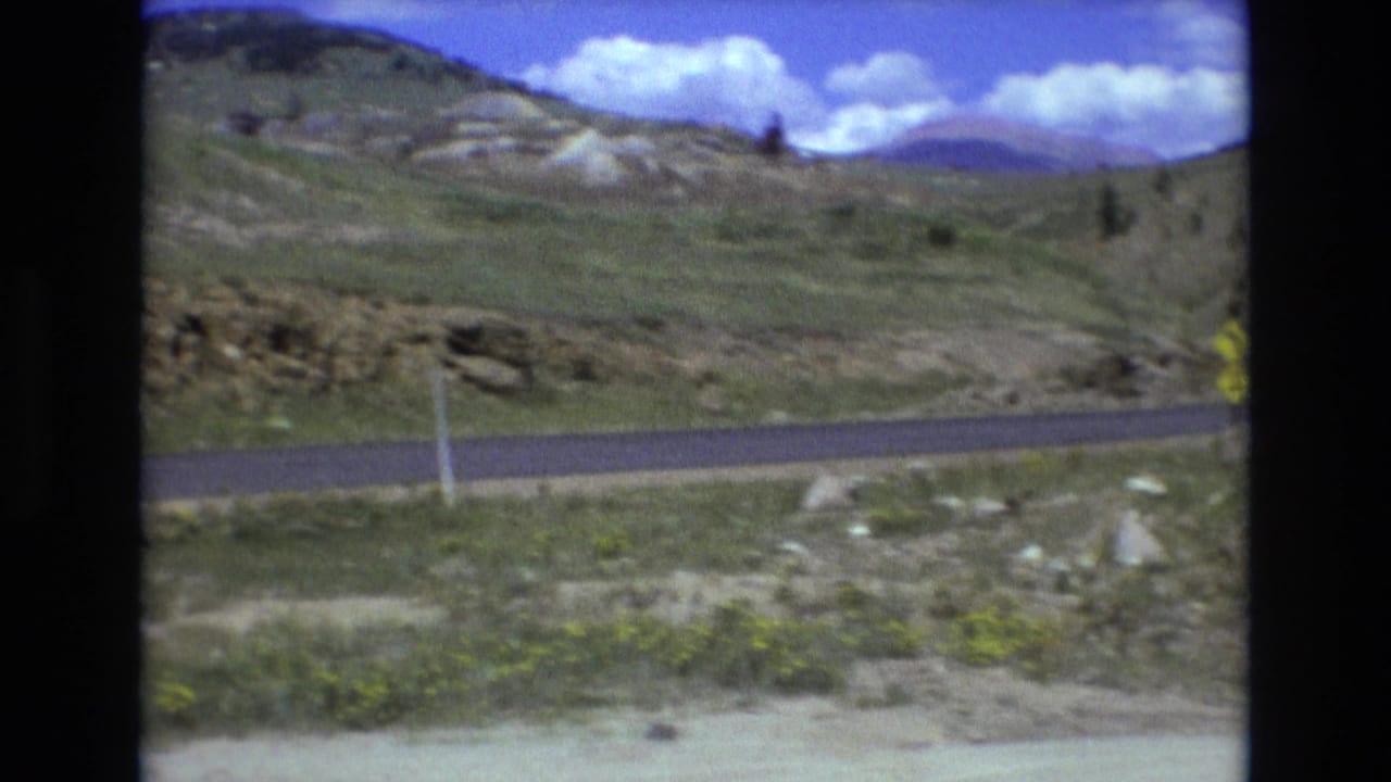 1973 8mm frame from the Colorado Springs area showing a paved road crossing the lower edge of rocky grassy hills beneath a saturated blue sky with white clouds.