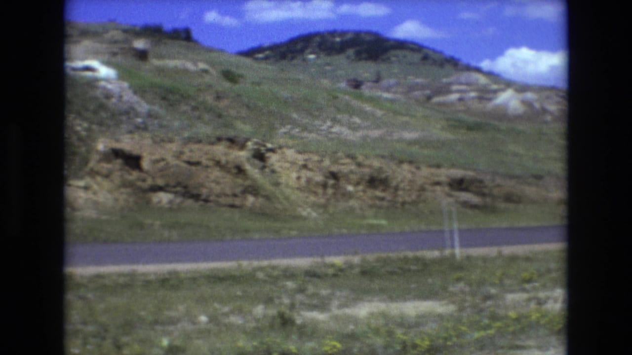 Roadside slope with rock ledges, grass, and distant hills under clouds.
