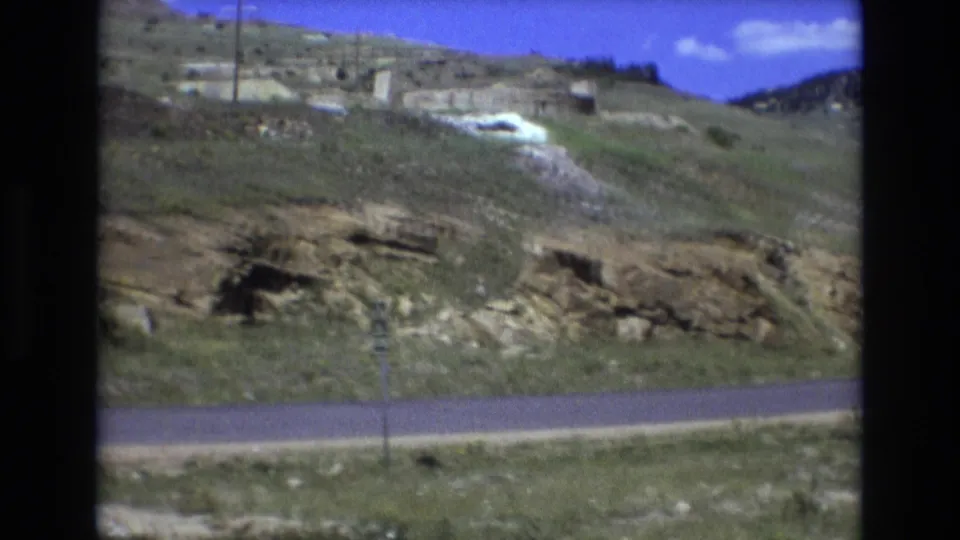 1973 8mm Colorado Springs-area road view with a rocky hillside, sparse vegetation, utility poles, and small pale structures or roadside features on the slope.