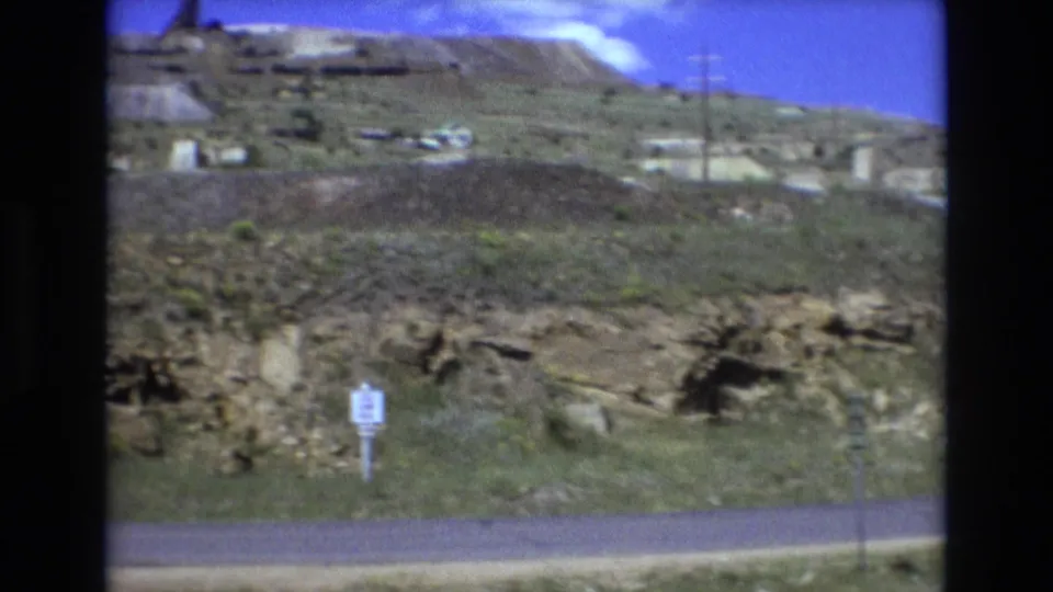 1973 8mm frame from the Colorado Springs area showing a small roadside sign in front of rocky grassland, with utility poles and pale slope features in the background.