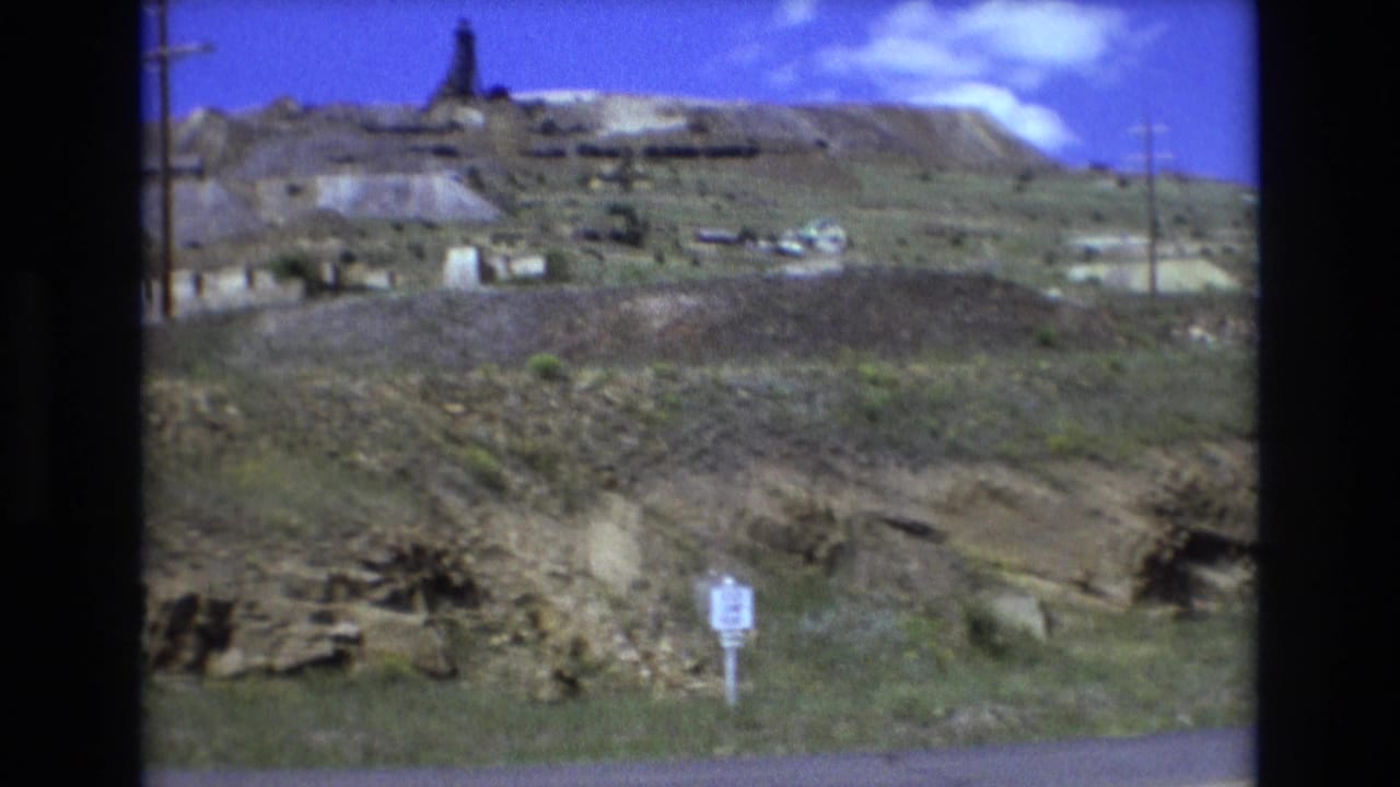1973 Colorado Springs 8mm frame showing a dry excavated hillside with low structures, a small foreground sign, utility poles, and bright blue sky.
