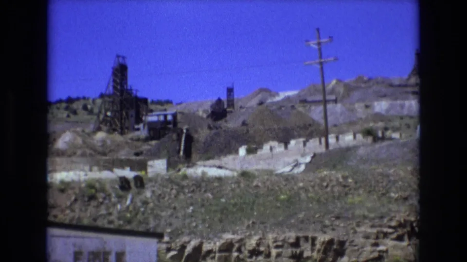 1973 Colorado Springs 8mm frame with a dark derrick-like structure, graded hillside, utility pole, low wall or buildings, rough foreground earth, and blue sky.