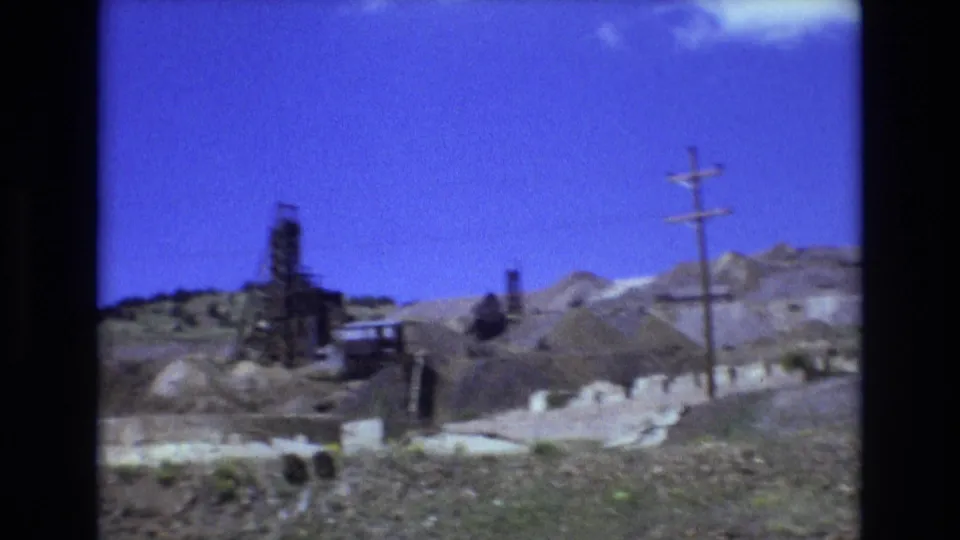 1973 Colorado Springs frame showing a roadside view of an industrial hillside with a derrick-like structure, utility pole, exposed earth, low structures, and open blue sky.